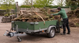Man laadt groenafval met takken en hout in een groene aanhanger op een tuincentrumterrein.